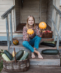 Girl is sitting on steps of an old village house with pumpkin in hands near baskets with zucchini and apples. Harvest. Autumn. Fall. Halloween. Thanksgiving day