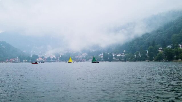 small colorful sailboat with people sitting on it passing row boats and pedal boats on a foggy day with buildings in the background on the lake of Nainital in uttarakhand