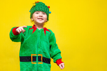little girl in elf costume and Christmas tree-shaped hat gallops on yellow background points finger...