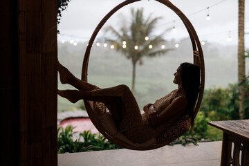 Woman in a rattan chair in a bamboo house enjoys a tropical holiday in Bali. Relax on the outdoor...