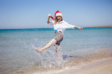 A cheerful young woman in a santa cap runs along the sea or ocean, splashing water