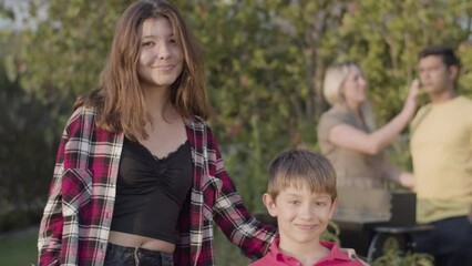 Happy teenage girl standing with her preteen brother, looking at camera and smiling outdoors, their parents standing at barbecue grill in background. Family at barbecue party concept