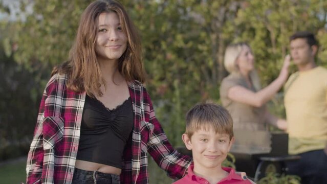 Happy teenage girl standing with her preteen brother, looking at camera and smiling outdoors, their parents standing at barbecue grill in background. Family at barbecue party concept