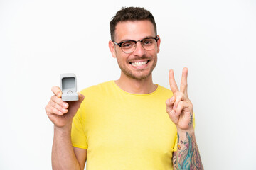 Young caucasian man holding a engagement ring isolated on white background smiling and showing victory sign