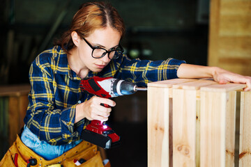 Woman in glasses using drill while working in carpentry workshop.