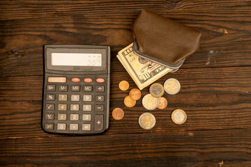Dollar banknotes, euro coins, a vintage brown leather wallet and a calculator on a wooden surface.
