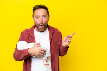 Middle age caucasian man holding his newborn son isolated on yellow background surprised and pointing side