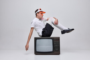 Portrait of stylish young man, boy posing in white tights and blac shirt, sitting on retro TV isolated over grey background