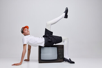 Portrait of stylish young man, boy posing on retro TV isolated over grey background. Techno shop...