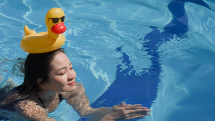 A cute attractive female person swimming in a swimming pool with a funny inflatable yellow duck on head. Asian girl smiling happily as she is bathing in blue water at luxury spa hotel. Soft focus.