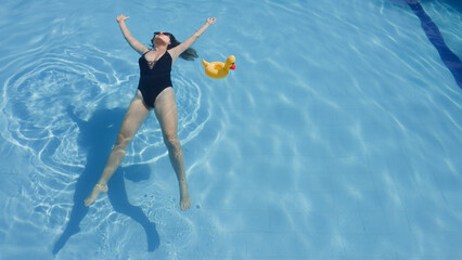 Woman wearing black swimsuit with yellow inflatable duck laying on surface of swimming pool. Girl with white skin floating on water with texture of ripples and sundog outdoors in summer. Soft focus.
