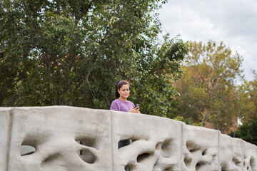 beautiful woman in purple blouse smiling in a park looking at her cell phone on a white bridge