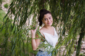 Smiling woman wearing white and denim skirt under weeping willow tree in park looking at camera seen from above.