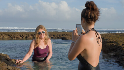 Happy family on holiday at ocean shore. Adult woman taking picture of senior woman as they are standing in sea water inside reef pool. Happy time on beach in summer. Soft focus.
