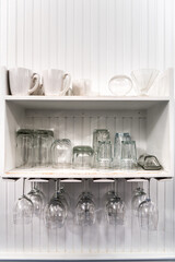 Closeup of modern kitchen room interior design white cabinet shelves with white cups, upside down hanging empty wine glasses rack for storage