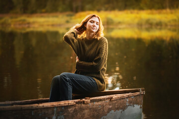 A beautiful blonde woman is sitting on the edge of an old fishing boat floating in a lake in the...