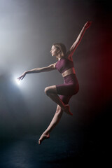 Young sporty and beautiful female woman jumping in fog/ smoke. Girl wearing fitness dance clothing making dance element performance on isolated black background with red light.
