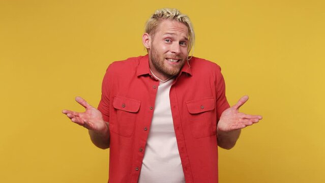 Young fun confused shy shamed blond man with dreadlocks 30s he wear red shirt white t-shirt look camera spreading hands say oops ouch oh omg i am so sorry isolated on plain yellow background studio
