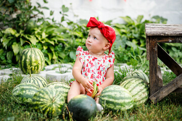 Fototapeta premium Portrait of a girl looking up in surprise in a red headband sitting near watermelons at the fair. Fresh ripe green watermelons on the grass on a sunny summer day