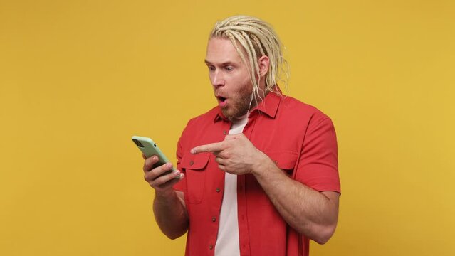 Young blond man with dreadlocks wear red shirt white t-shirt hold use mobile cell phone typing browsing swipe chat send sms do online shopping order delivery home isolated on plain yellow background