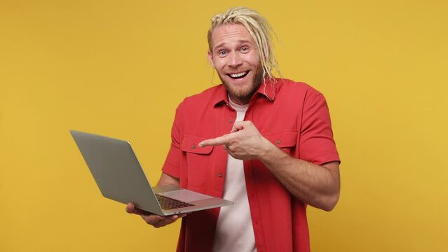 Young surprised shocked happy blond man with dreadlocks 30s he wear red shirt white t-shirt hold in hands typing use work point index finger on laptop pc computer isolated on plain yellow background