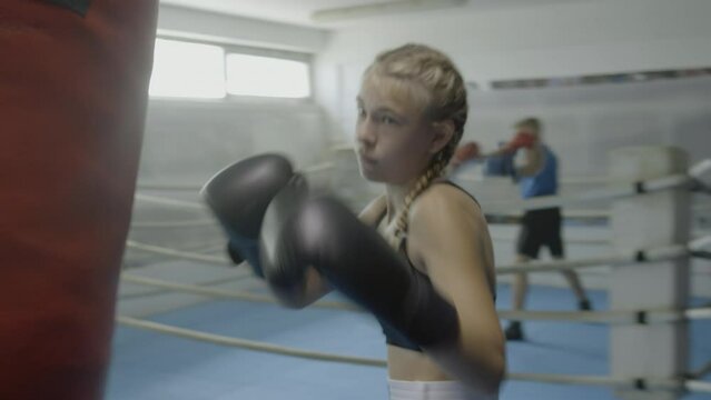 Teen girl training with punchbag, boxing class workout, handheld shot. Boys sparring in ring, teenager practicing kicks with equipment in gym