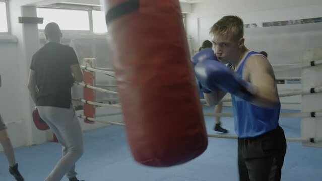 Teen boy training with boxing pear at fighting class workout, handheld shot. Teenagers sparring in ring in background, practicing kicks in gym