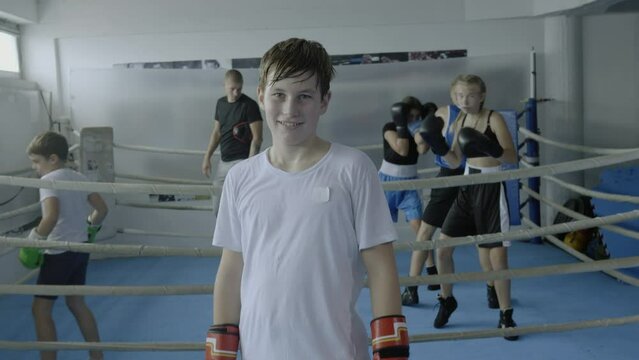 Boy holding hands in boxing gloves near face, looking at camera, zoom out shot. Child exercising in gym portrait, practicing at kid fighting class