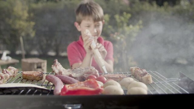 Happy preteen boy taking meat with fork from grill to eat it. Boy standing at barbecue grill with roasted pork, kebab, pepper and potatoes outdoors. Barbecue party concept
