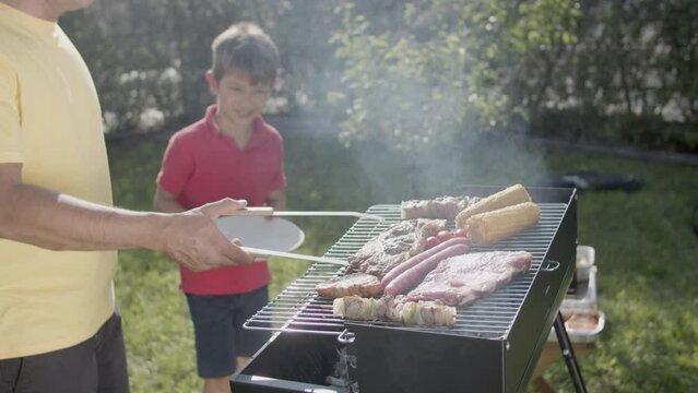 Man putting grilled pork ribs with tongs on plate of his smiling son. Man cooking roasted meat, corn and cherry tomatoes on barbecue grill. Barbecue party and family leisure concept