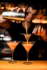 hand of bartender holds shaker with strainer and accurate pours cocktail into martini glass.