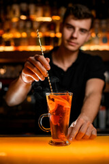 hand of bartender holds spoon and stirs drink with lemon in transparent cup on the bar