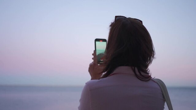 Woman traveler taking picture of the sea at sunset on the smartphone camera. Back view portrait mode slow motion shoot of adult woman filming video seascape on phone camera