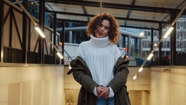 Portrait pretty cheerful curly haired woman posing in city in evening looking at camera smiling seductively, relaxed female backlit with warm street lights, urban style, autumn outfit pullover coat