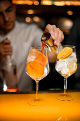 Close-up of glass with ice cubes and orange slices on the bar and bartender pours alcoholic drink from jigger into glass