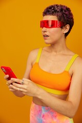 Woman blogger surprise in unusual millennial glasses taking selfies on her phone sporting brightly colored clothes against an orange studio background, free space