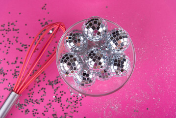 Christmas, New Year party decoration. Shining silver disco balls lay in transparent glass plate, kitchen whisk on bright pink background. Small shiny stars, glitter around. Top view, flat lay, mirror