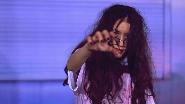 Beautiful caucasian teenage girl with flowing long hair and a bloody dress stretching her hand to the camera walks like a zombie against a blue wall, having fun at a school party, celebrating Hallowee