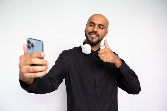 Portrait of happy young man taking selfie with mobile phone against white background. Bearded businessman wearing black shirt and headphones making thumb up and smiling. Social networking concept