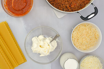 Raw lasagna noodles, ground beef on frying pan, marinara, cream cheese, mozzarella, parmesan cheese, milk. Step by step beef lasagna recipe, ingredients on kitchen table