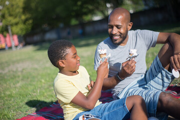 Naklejka na ściany i meble Happy daddy and little son spending time together. Smiling African American man and cute boy sitting on ground holding ice-cream cones talking in summer park. Parenting and leisure concept