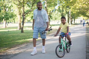Portrait of happy dad and son having good time in park. Handsome African American man with skateboard in his hand walking looking at camera and nice boy riding bike. Leisure and childhood concept