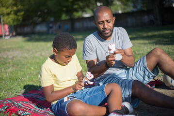 Naklejka na ściany i meble Portrait of happy man and boy resting together on ground. African American father and his kid sitting on blanket in summer park concentrated on unwrapping ice cream cones. Resting and leisure concept
