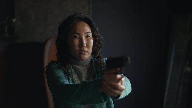 4k Young woman holds gun and stands in retro interior spbd. Killing eve concept. Beautiful Asian female is holding weapon and aiming, showing confusion for camera and standing in dark room. One