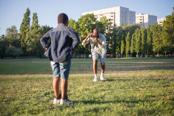 Portrait of dad and son taking photo on field. Handsome bearded man and boy standing on grassy ground, father with camera taking picture of kid. Hobby, leisure and having rest together concept