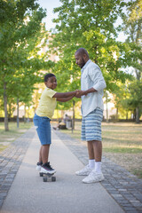 Side view of African American man and boy standing on shady alley. Serious father looking at little son holding his hands and kid on skateboard looking at camera. Summer activity, healthy rest concept