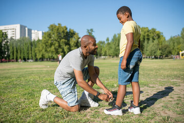 Side view of African American dad and his son standing on field.Man standing on his one knee on grass tying shoelaces on kid sneakers both looking at each other.Parents care and sport activity concept