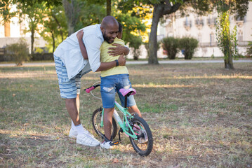 Portrait of happy African American dad and son training in park. Smiling man and boy with bike standing in city park and father hugging kid with love. Active summer leisure and parenthood concept