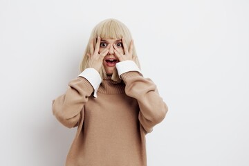 Horizontal photo, a woman on a white background in a beige sweater with beautiful blond hair hides her face behind her palms with fingers spread