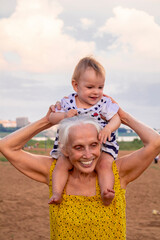 The kid sits on the shoulders of his grandmother on the outdoors beach. Clothes in bright yellow, blue and white. Baby and grandma are smiling.